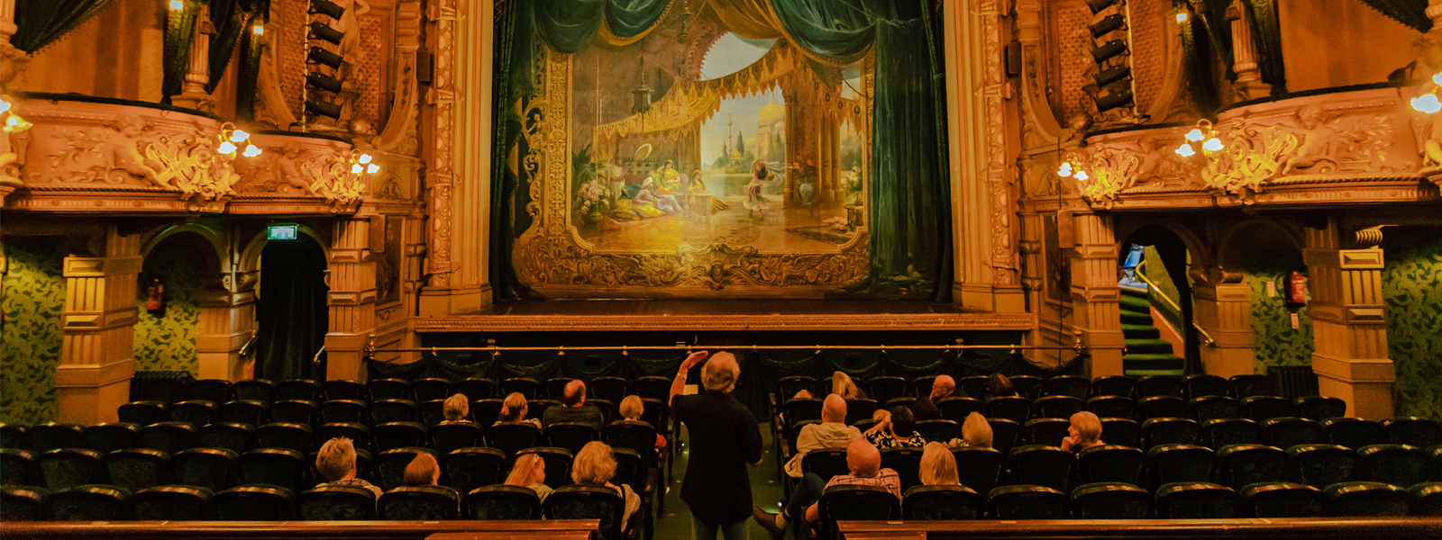 A group of visitors sat in the Isle of Man's prestigious Gaiety Theatre on a guided tour, the tour guide is stood in the aisle explaining the history of this grand venue.